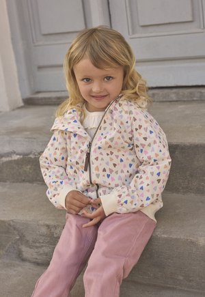 Lichte, licht crèmekleurige jas met een hartjespatroon, voorzien van een ritssluiting aan de voorkant en elastische manchetten, gedragen over een wit shirt en gecombineerd met roze broek.