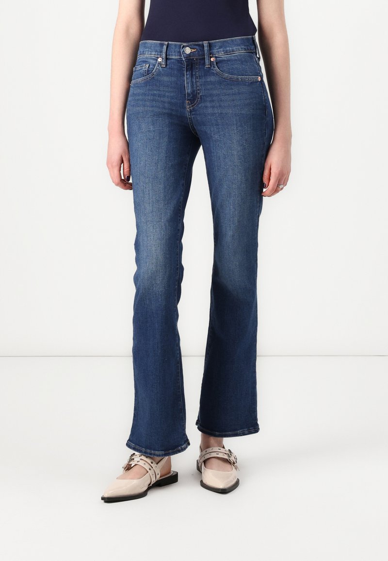 Jean en denim bleu foncé avec une coupe droite, cinq poches et des détails de couture subtils aux coutures. Associé à des chaussures plates beiges.