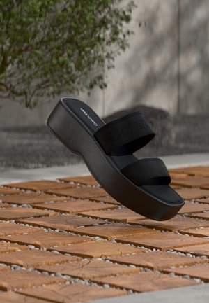 Μαύρο πλατφόρμα σανδάλι slide Calvin Klein με δύο φαρδιές λουρίδες που αιωρούνται πάνω από επιφάνεια από καφέ ξύλο με πλακάκια σε εξωτερικό χώρο.