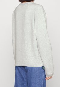 Svetle šedá mikina s volným střihem, měkkou texturou, spadlými rameny a šňůrkou na spodním lemu, kombinovaná s modrými džíny.