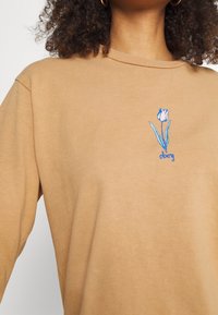 Brun bomullströja med rund halsringning, som har en broderad blå och vit tulpan på vänster bröst.