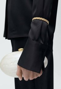 Cămașă neagră din satin cu manșete largi, având o bandă decorativă aurie. Mână care ține o geantă albă glossy, în formă ovală, cu un capac auriu.