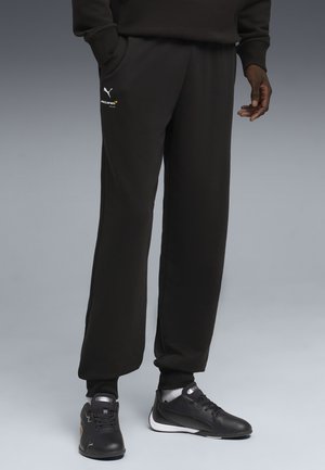 Homme portant un pantalon de survêtement de course McLaren noir et des baskets noires Puma, debout devant un fond gris.