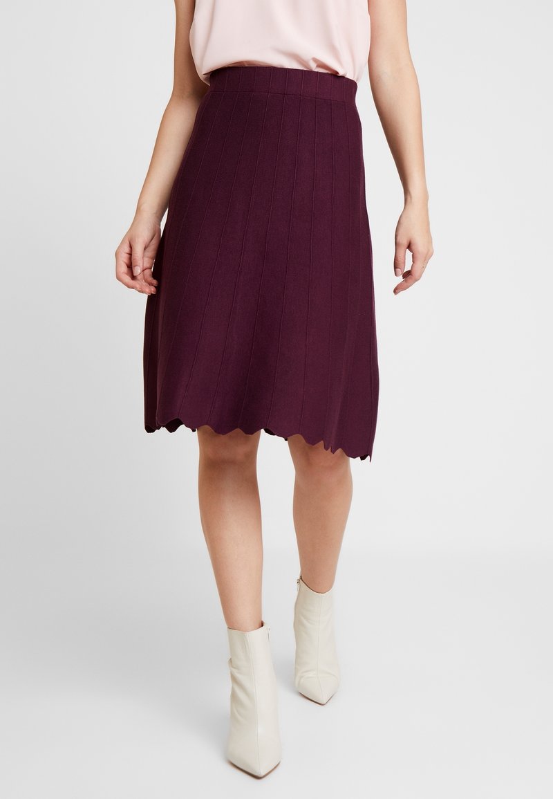 Jupe bourgogne midi avec une texture côtelée et un ourlet à volants, associée à des bottes blanches à la cheville, sur un fond uni.