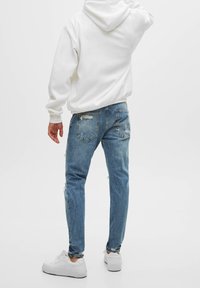Man som bär en vit hoodie, slitna blåjeans och vita sneakers står mot en vit bakgrund, vänd bort med en arm utsträckt.