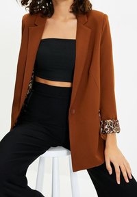 Mujer sentada en un taburete blanco, vestida con un blazer naranja quemado con puños de estampado de leopardo, un top tube negro cortado y pantalones negros de cintura alta.