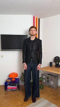 Black textured blazer over a black shirt, paired with wide-leg blue jeans and burgundy shoes. Features tattoos on arms and minimal accessories.