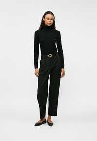 Fekete garbó pulóver, szabott fekete nadrág zsebekkel és fekete lapos cipő. Az öltözetet egy aranycsatos öv és letisztult forma jellemzi.