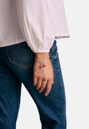 Silbernes Gliederarmband mit roten Anhängern, getragen an einer Hand. Die Person trägt ein weißes Oberteil und blaue Jeans.