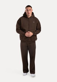 Brązowy hoodie na zamek błyskawiczny z kapturem na sznurku, w zestawie z dopasowanymi brązowymi spodniami. Zawiera mały detal z logo i ściągacze na rękawach.