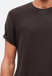 Horná časť tela osoby v tmavo hnedom tričku s krátkym rukávom a zvinutými rukávmi na jednofarebnom pozadí.