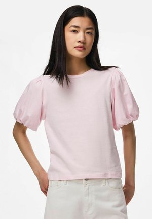 Jeune femme aux cheveux raides et foncés portant une chemise rose pâle à manches bouffantes et un pantalon blanc sur un fond uni.
