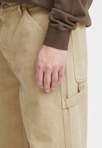 Beige pologamasiner med lige snit, der har sidelommer og en subtil tekstur. En brun sweater er synlig ved håndleddet.