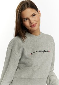 Grijze sweatshirt met een geribbelde ronde hals, lange mouwen en zwart geborduurde tekst met roze hartaccenten.