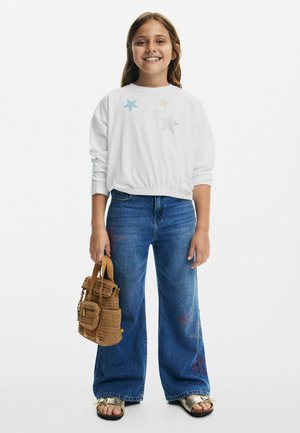 Vit långärmad sweatshirt med stjärndekorationer, i kombination med vida blå jeans och en flätad brun handväska. Sandaler med en metallisk rem.