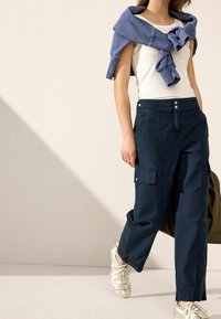 Marine cargobroek met voor- en zijzakken, voorzien van zilveren drukknoopsluiting, gecombineerd met een witte tanktop en een blauwe sweatshirt nonchalant over de schouders gedrapeerd.