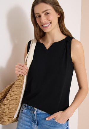 Nainen hymyilee, pukeutuneena mustaan hihattomaan toppiin ja sinisiin farkkuihin, kantaen kudottua beigeä olkalaukkua olallaan.