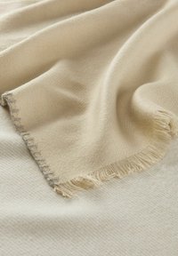 Couverture beige avec une texture douce, des bords effilochés et un motif tissé subtil, posée sur une surface plus claire avec un motif.