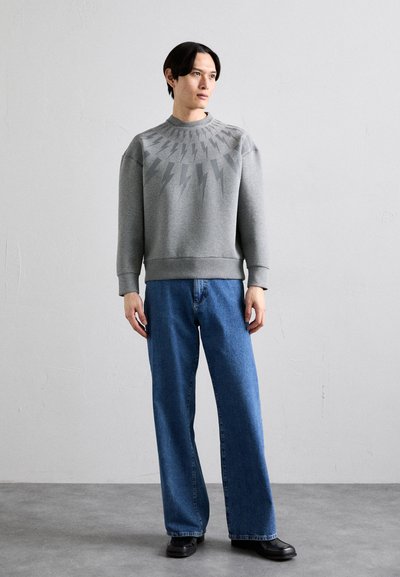 Grå sweatshirt med blixtmönster vid halsringningen, tillsammans med blå vida jeans och svarta ankelkängor. Slät tyg, avslappnad passform.