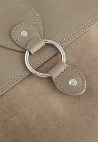 Beežist nahast käekott tekstuurse viimistlusega, millel on hõbedane metallist rõngas kinnitatud rihmale, mis sulgub klambriga, ning kontrastsed õmblused.