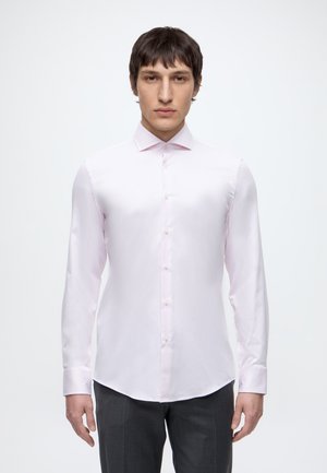 Jeune homme aux cheveux courts et foncés portant une chemise rose clair à manches longues boutonnée et un pantalon foncé sur un fond blanc uni.
