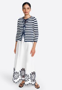 Knitted navy and white striped cardigan with 3/4 sleeves, paired with a white maxi skirt featuring black floral cut-out designs. Flat sandals.