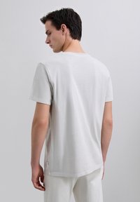Uomo con capelli scuri che indossa una maglietta grigia chiara a maniche corte e pantaloni bianchi, visto di spalle su uno sfondo neutro.