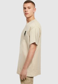 Beige bomulds t-shirt med rund hals, korte ærmer og en lille sort grafik på brystet. Lige pasform og blød tekstur.