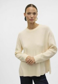 Beige ribbet sweater med rund halsudskæring, faldne skuldre og sideslids, parret med mørke jeans. Glat tekstur, afslappet pasform.