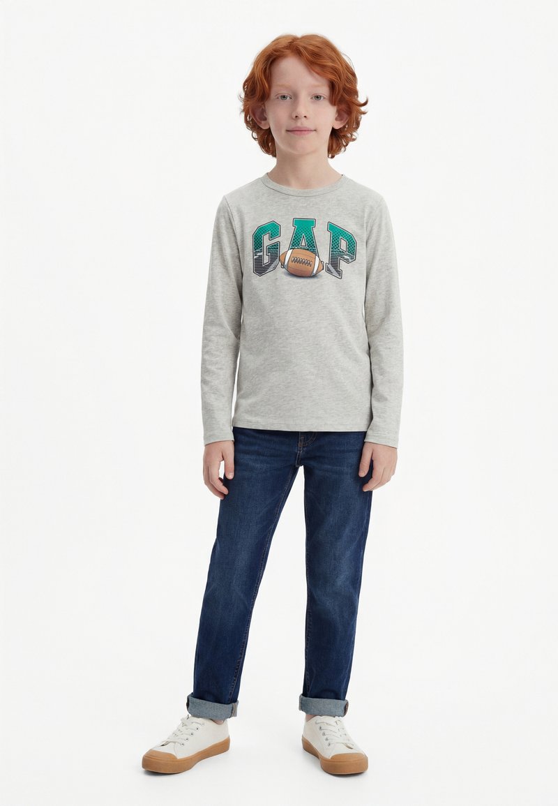 Παιδί με κόκκινα μαλλιά στέκεται φορώντας γκρι μπλουζάκι μακρυμάνικο με το γράμμα GAP και γραφικό ποδοσφαίρου, σκούρο μπλε τζιν και λευκά αθλητικά παπούτσια με καφέ σόλες.