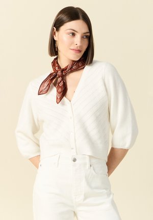 Kobieta o prostych brązowych włosach, ubrana w białą rozpinaną kardigan i spodnie oraz brązową apaszkę z wzorem zawiązaną na szyi, patrząca na bok.