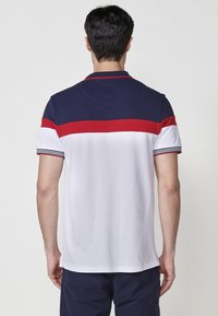 Polo majica z navy modrim zgornjim delom, rdečo horizontalno črto in belim spodnjim delom. Kratki rokavi z črtastimi manšetami. Gladka tekstura tkanine.