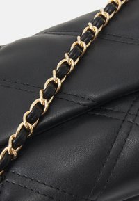 Musta nahkainen käsilaukku, jossa on tikattu kuvio ja kultainen sekä musta punottu ketjuhihna. Sileä pinta ja tarkat saumat.