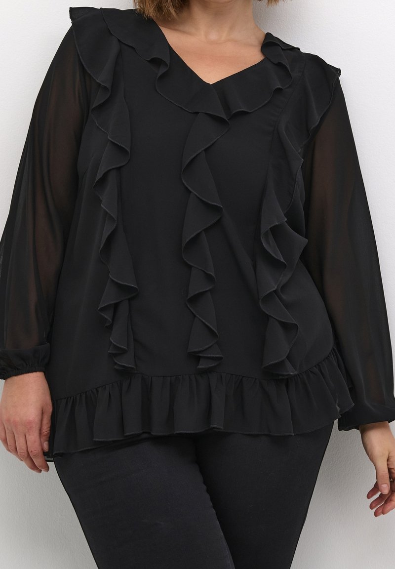 Femme portant un chemisier noir à manches longues avec bordure à volants et manches transparentes, associé à un pantalon noir, debout sur un fond blanc.