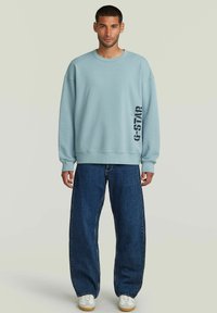 Sudadera azul claro con puños y dobladillo acanalados, con un logo impreso en negro en el lado izquierdo, combinada con unos jeans oscuros de corte holgado.