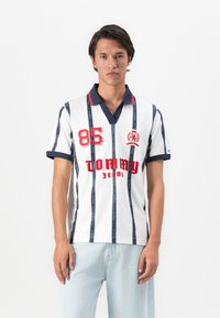 Jonge man in een wit poloshirt met marineblauwe strepen, rode "85" en "Tommy Jeans" tekst, gecombineerd met lichtblauwe jeans tegen een effen achtergrond.