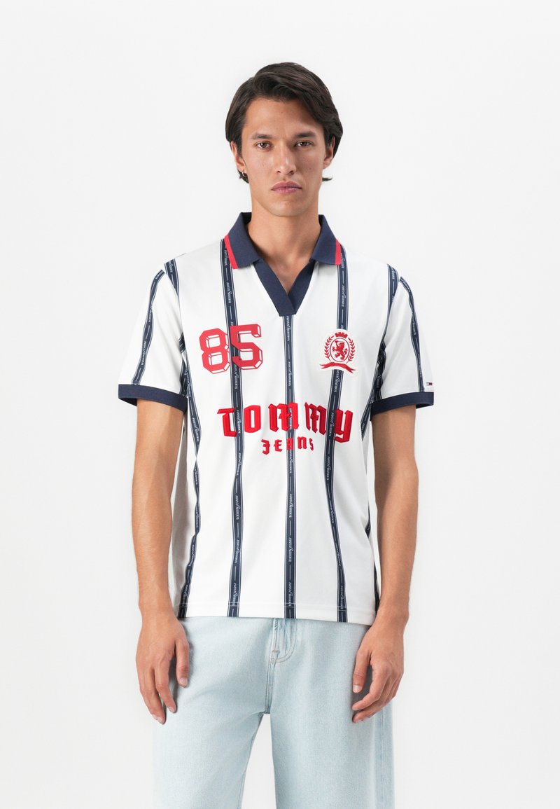 Jonge man in een wit poloshirt met marineblauwe strepen, rode "85" en "Tommy Jeans" tekst, gecombineerd met lichtblauwe jeans tegen een effen achtergrond.