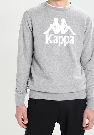 Grauer Sweatshirt mit weißem Kappa-Logo, das zwei Rücken-an-Rücken sitzende Figuren oberhalb des Wortes "Kappa" zeigt, getragen von einer Person in schwarzer Hose.