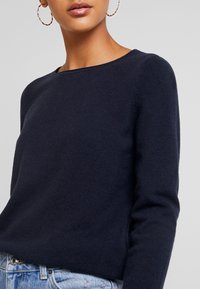 Pull bleu marine à col rond, à manches longues et à texture douce. Porté avec un jean en denim clair, présentant un subtil détail côtelé à l'ourlet.