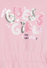 Rosa Baumwoll-T-Shirt mit mehrfarbigem Pailletten-Text: "HAPPY GUESS GIRL ON VACATION", versehen mit Herzen und dekorativen Akzenten.