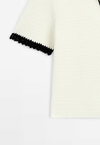 Haut à manches courtes blanc en crochet avec motif texturé et bordure noire festonnée aux extrémités des manches et à l'encolure.