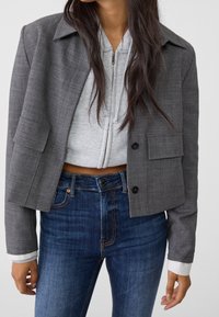 Mujer vistiendo una chaqueta gris corta sobre una sudadera con cremallera de color gris claro, combinada con jeans azules de tiro alto, mostrada de cuello a caderas.