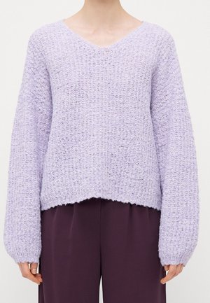 Personne portant un pull en maille texturée violet clair à manches longues et col en V, associé à un pantalon violet foncé.