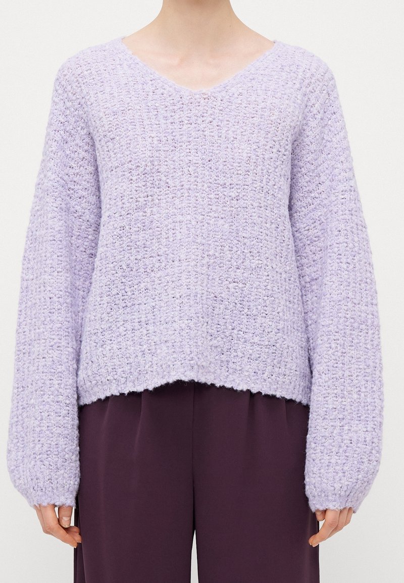 Personne portant un pull en maille texturée violet clair à manches longues et col en V, associé à un pantalon violet foncé.