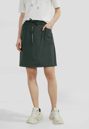 Femme portant une jupe noire à cordon avec de grandes poches avant, des baskets blanches de sport et un haut blanc à manches courtes.