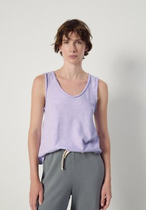 Woman with short brown hair wearing a sleeveless lavender top and gray drawstring pants, standing against a plain light background.