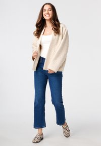 Beige gewatteerde oversized cardigan, witte tanktop, blauwe rechte jeans en gestreepte instappers met een lichte achtergrond.