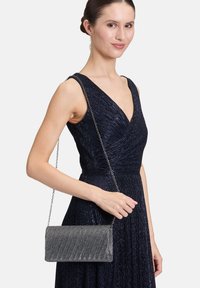 Pochette en métal argenté avec un motif texturé en rayures diagonales, dotée d'une chaîne, tenue par un mannequin portant une robe sombre.