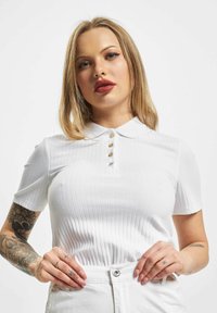 Hvit ribbestrikket polotrøye med korte ermer, krage og tre dekorative knapper på stolpen. Glatt tekstur.