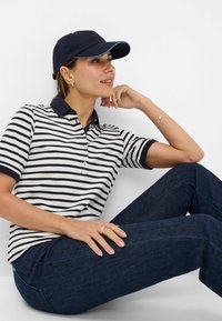 Polo a righe in crema e blu navy con maniche corte, abbinato a jeans in denim scuro e a un cappellino blu navy con un accento azzurro.